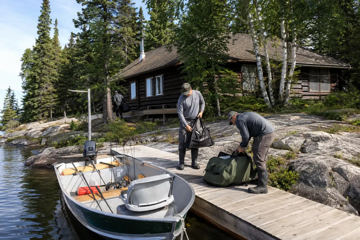 Anglers unload fishing gear beside a remote cabin, dock and aluminum boat.